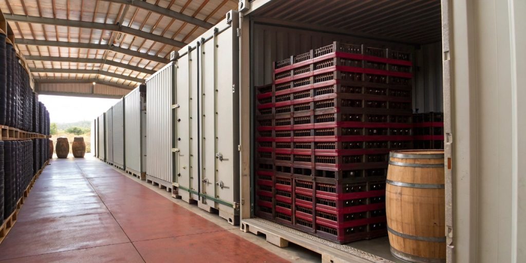 Commercial wine storage facility showing shipping containers, stacked wine bottle crates in red plastic cases, and wooden barrels prepared for bulk wine shipping operations in an industrial warehouse setting with concrete floors and steel beam construction.