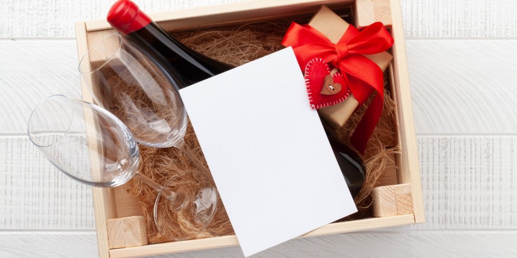 Complete wine gift box designed to send wine to a friend, featuring wine bottle with red cap, two wine glasses, blank greeting card, and small wrapped present with red ribbon and heart ornament nestled in natural straw packaging in wooden crate.