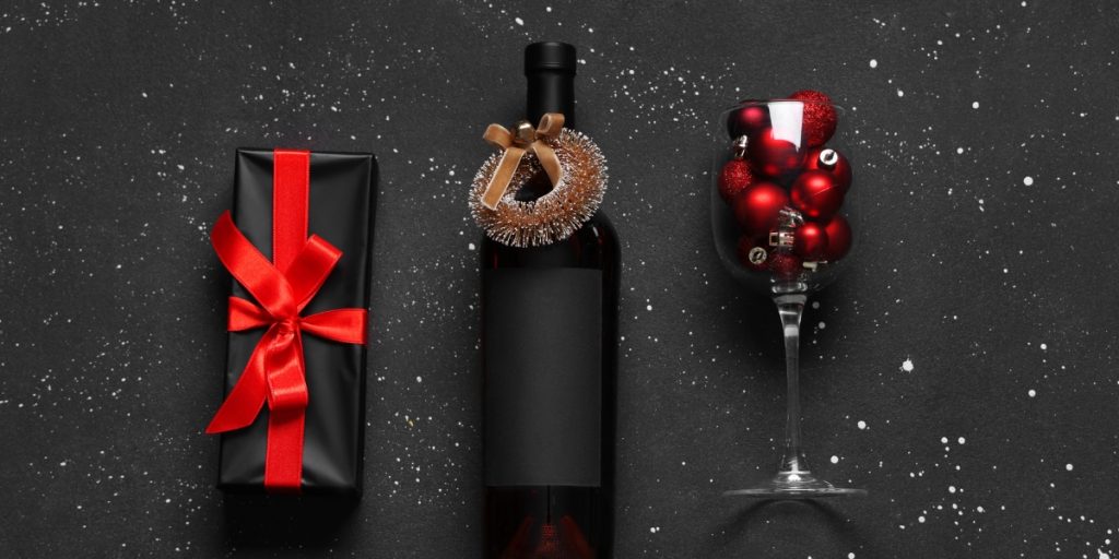 Elegant gift presentation showing how to send wine to a friend with black wrapped wine bottle decorated with miniature wreath, matching black gift box with red satin bow, and wine glass filled with red ornaments on snowy black background for holiday gifting.