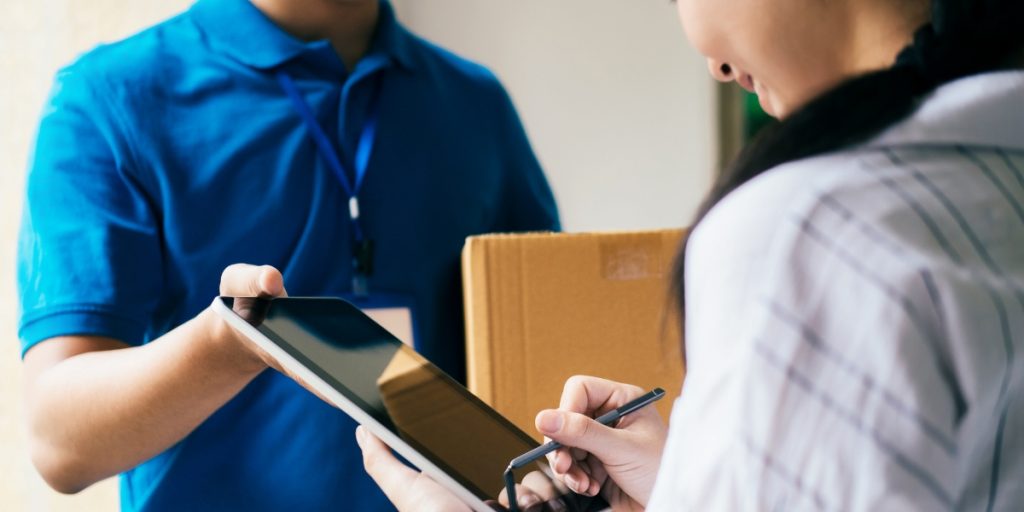 Delivery person in blue uniform presenting cardboard package to customer signing on digital tablet for shipping wine across country service confirmation at residential doorstep.