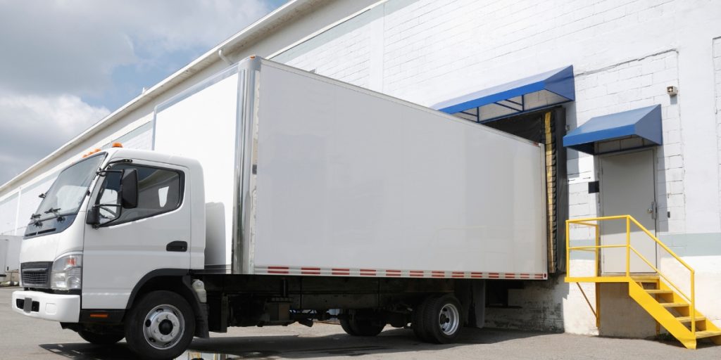White refrigerated delivery truck with insulated cargo box backed up to warehouse loading dock with yellow stairs, demonstrating temperature-controlled shipping logistics for perishable goods transportation.