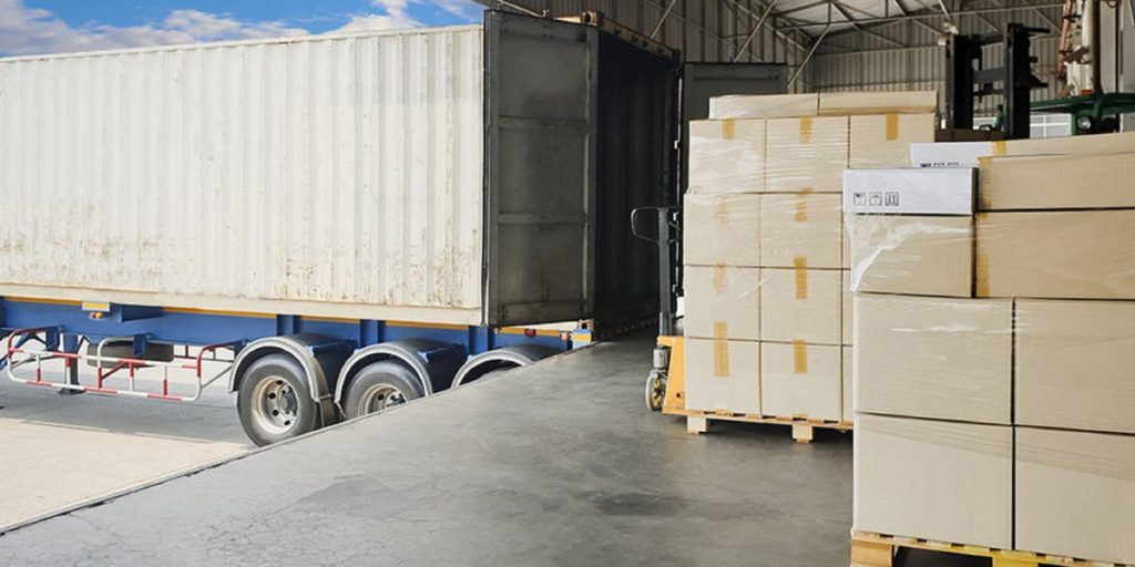 A long-distance wine shipping process shows cardboard boxes stacked on pallets and loaded into a commercial shipping container with a forklift at an industrial warehouse facility.