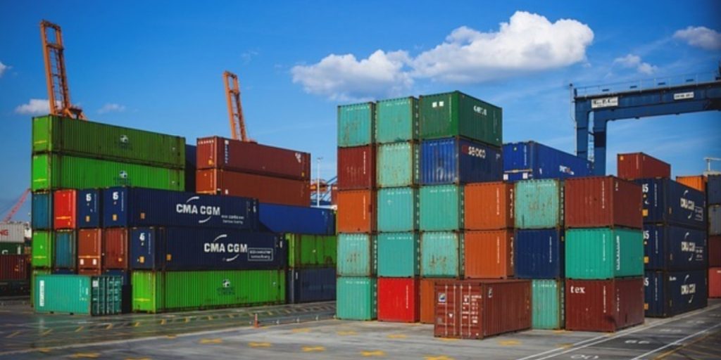 Stacked shipping containers in various colors at a port facility with cranes and loading equipment, representing the logistics involved in international wine shipping customs and tariffs.