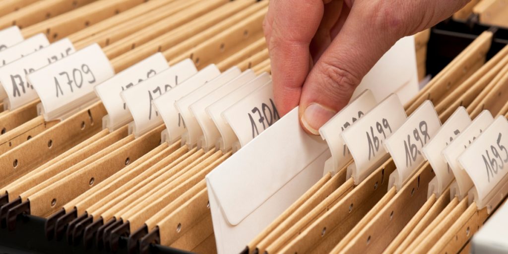 A person's hand reviews organized file folders with handwritten labels in a wooden filing system, representing documentation organization relevant to problems that delay wine shipments.