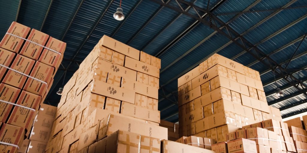 Outsourcing wine shipping vs in-house fulfillment showing warehouse with stacked cardboard boxes under industrial ceiling and lighting, representing storage and logistics infrastructure.