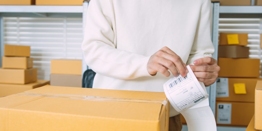 Outsourcing wine shipping vs in-house fulfillment depicting warehouse worker applying barcode label to cardboard package with organized boxes in background storage facility.