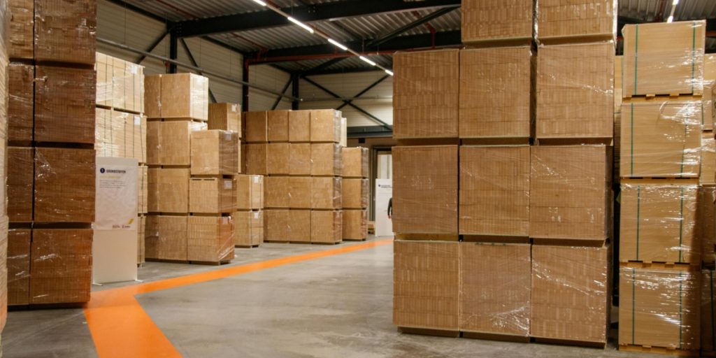 Outsourcing wine shipping vs in-house fulfillment showing organized warehouse with stacked pallets of cardboard boxes wrapped in plastic, orange floor markings, and industrial storage setup.