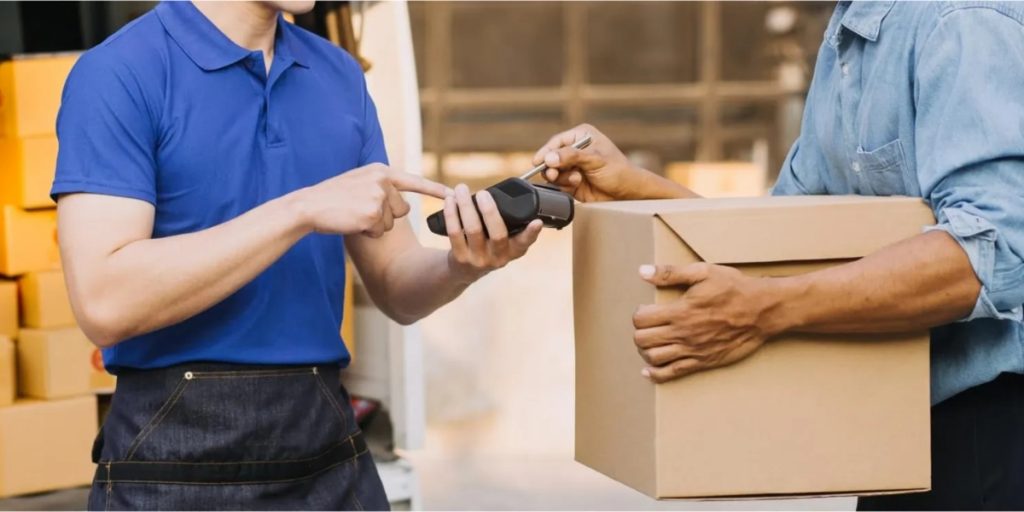 What states allow alcohol delivery showing a delivery driver in blue shirt processing a contactless payment with customer holding cardboard box package at warehouse with yellow bins.
