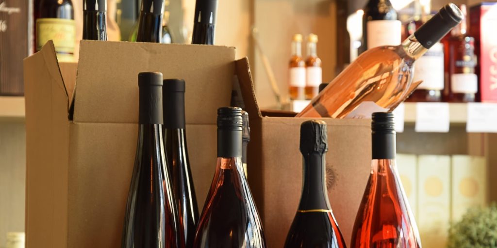 What states allow alcohol delivery illustration displaying wine bottles packed in cardboard shipping box with various red and rosé wines arranged for interstate or local delivery service.
