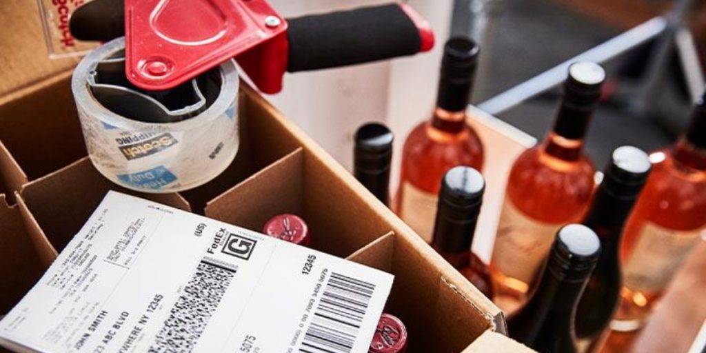 Photograph showing why USPS cannot ship wine, displaying wine bottles in a cardboard box with shipping labels, barcodes, and red wax seals indicating restricted alcohol shipment regulations.