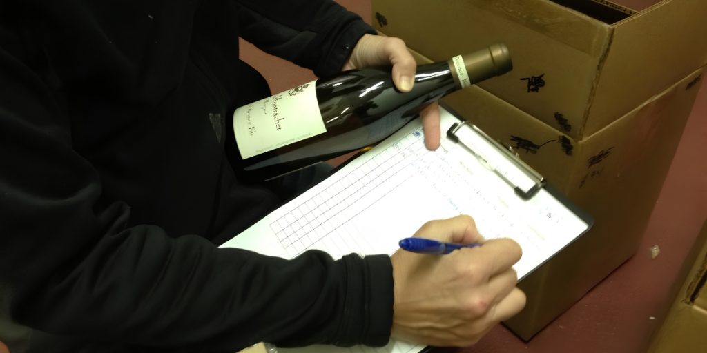 Wine club fulfillment: a worker holds a wine bottle and marks a clipboard during inventory documentation and shipment processing in a warehouse.