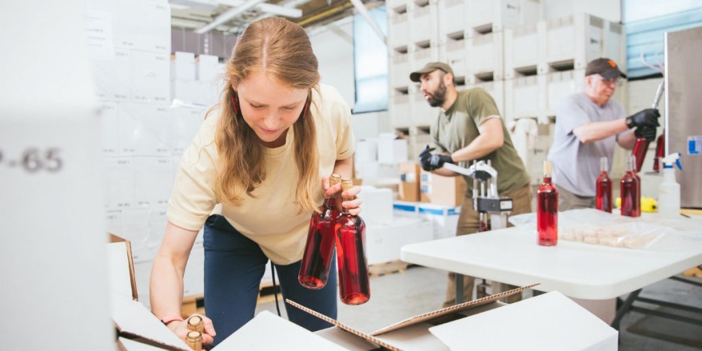 Wine club fulfillment: a woman in a yellow shirt inspects red wine bottles during quality control at a fulfillment facility with team members in the background.