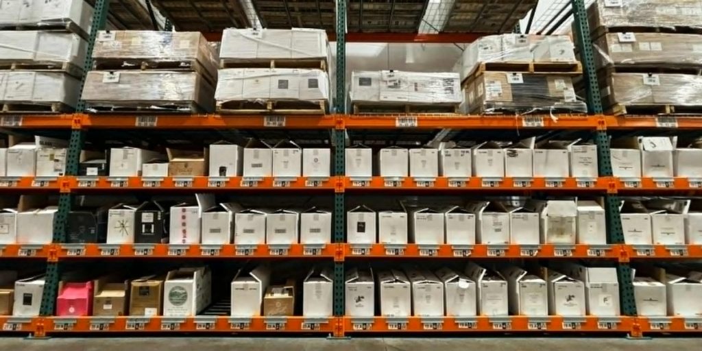 Wine club shipping - a warehouse storage facility with multiple shelves of packaged wine boxes and cases organized on orange and green industrial racking, showing inventory management and fulfillment operations for wine club distribution