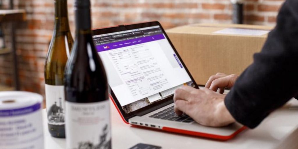 Wine club logistics featuring a staff member using a laptop to manage wine orders and shipment tracking with wine bottles displayed on a brick surface.