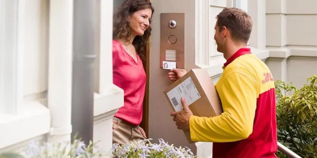 Adult signature wine delivery – delivery driver in yellow and red uniform presenting wine package to smiling woman at residential doorway, requiring adult signature for age-restricted beverage delivery