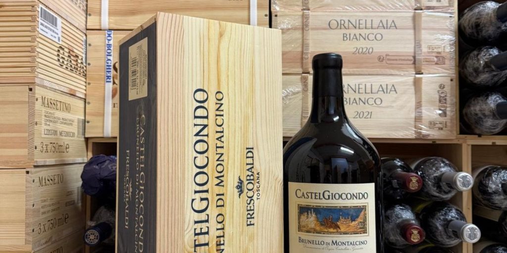 A large-format Castelgiocondo Brunello di Montalcino bottle stands beside its wooden case amid stacked crates, showcasing the appeal of large format wine bottles.