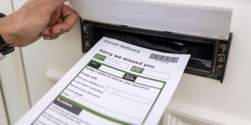 A hand pulls a “Sorry we missed you” parcel notice from a mail slot, detailing redelivery options after a missed wine delivery.